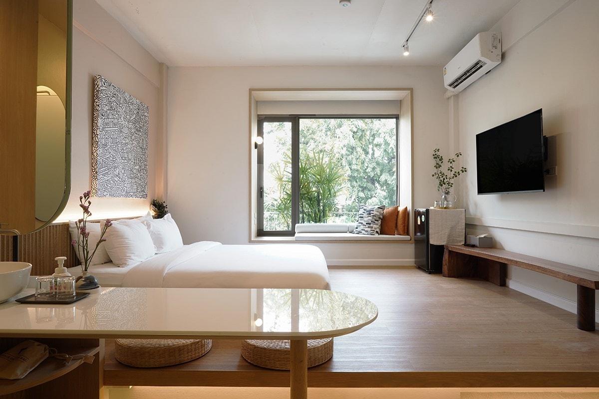 A spacious room features a premium king size bed and a window seat with views of greenery. A 50" smart TV is mounted on the wall. A wooden bench and a small table enhance the space, while natural light illuminates the room.