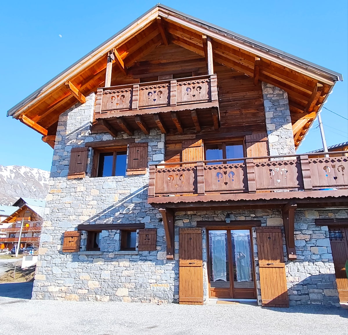 A traditional chalet features a stone facade with wooden accents, including balconies adorned with intricate carvings. Large windows provide views of the surrounding landscape. The entrance showcases double wooden doors, leading to a welcoming atmosphere in the Alps.