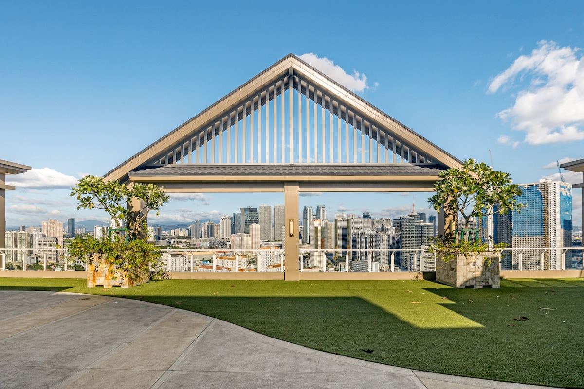 An expansive outdoor sky lounge features contemporary architecture with a triangular roof overhead. Lush planters house greenery, providing a touch of nature against the backdrop of a vibrant city skyline. Clear skies punctuate the serene atmosphere, enhancing the panoramic view.