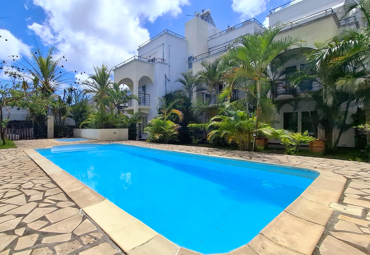 A communal swimming pool is framed by lush greenery and tropical plants. The clear blue water reflects sunlight, while the surrounding stone-paved area provides a welcoming space for relaxation. The two-story building's architecture features arched balconies, enhancing the serene ambiance of the pool area.