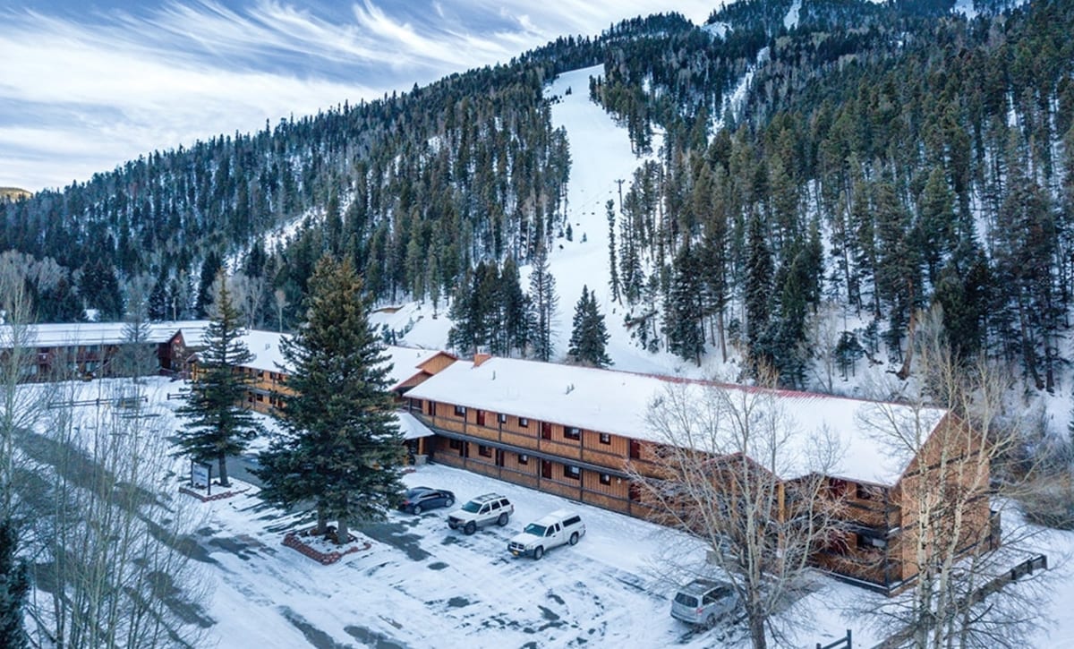 A snow-covered resort building is nestled against a backdrop of pine trees and snow-capped mountains. A ski slope is visible in the distance, enhancing the winter landscape. Several vehicles are parked in front of the property, and the ground is blanketed in fresh snow.