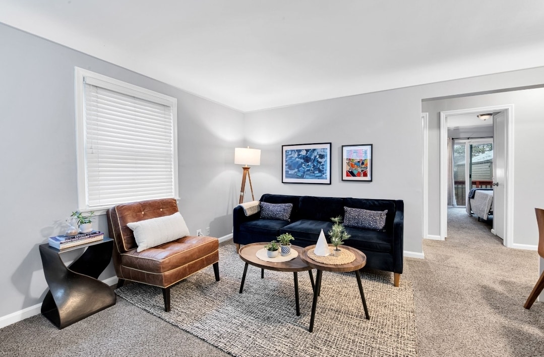 A living space is depicted, featuring a dark sofa and a mid-century armchair positioned around two round tables. Natural light fills the room through a nearby window, highlighting framed artwork on the gray wall. A plush area rug anchors the seating arrangement, creating a comfortable atmosphere.