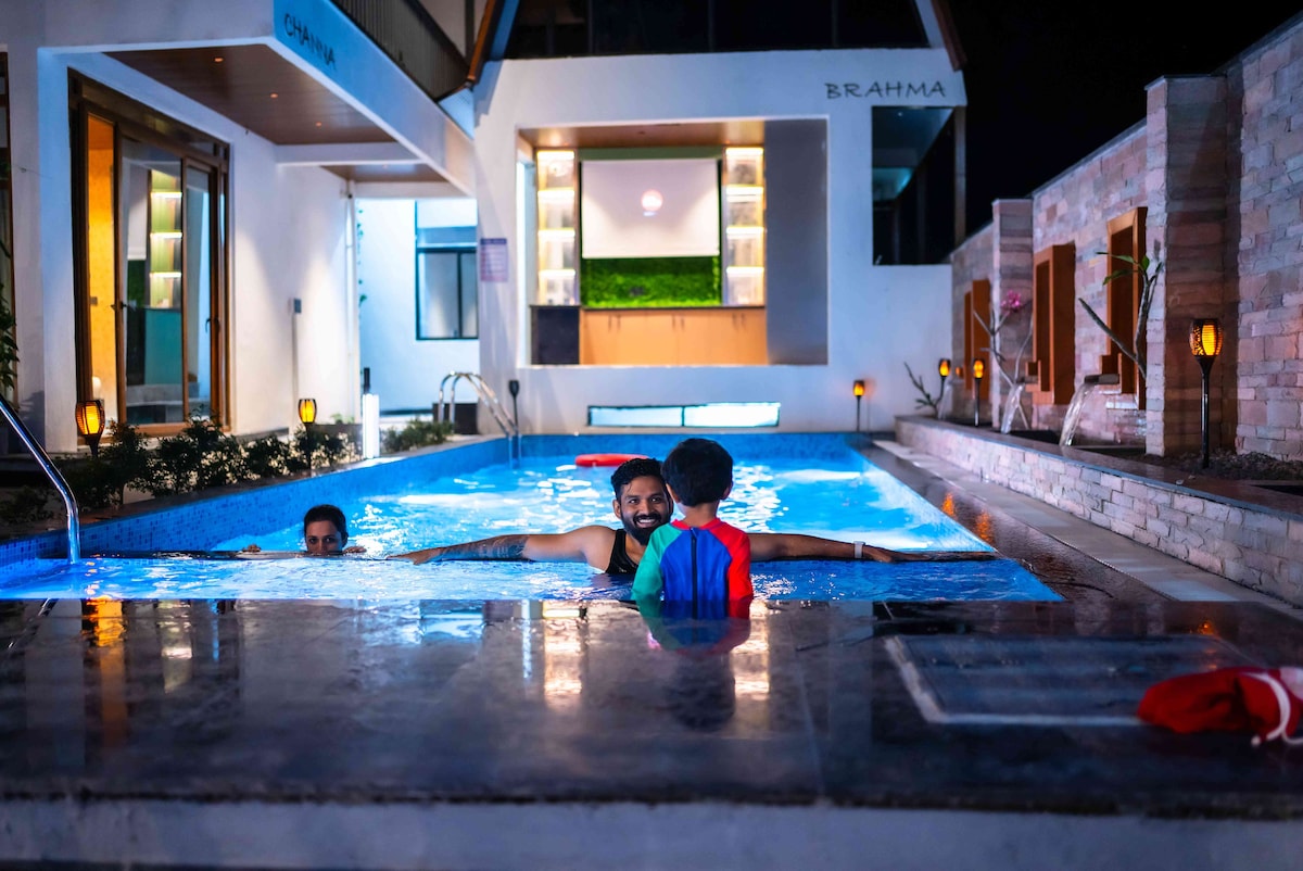A serene pool area is illuminated under soft lighting at night. The swimming pool is partially visible with a smooth stone edge, while a private cabana and seating area can be seen in the background. Warm light from lanterns highlights the tranquil atmosphere.