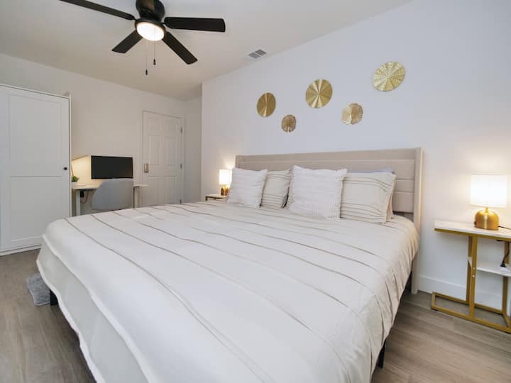 Dormitorio luminoso y espacioso con una cómoda cama tamaño king, un espacio de trabajo dedicado y ventilador de techo para enfriar. La paleta de colores neutros y los detalles dorados crean un ambiente relajante y elegante.