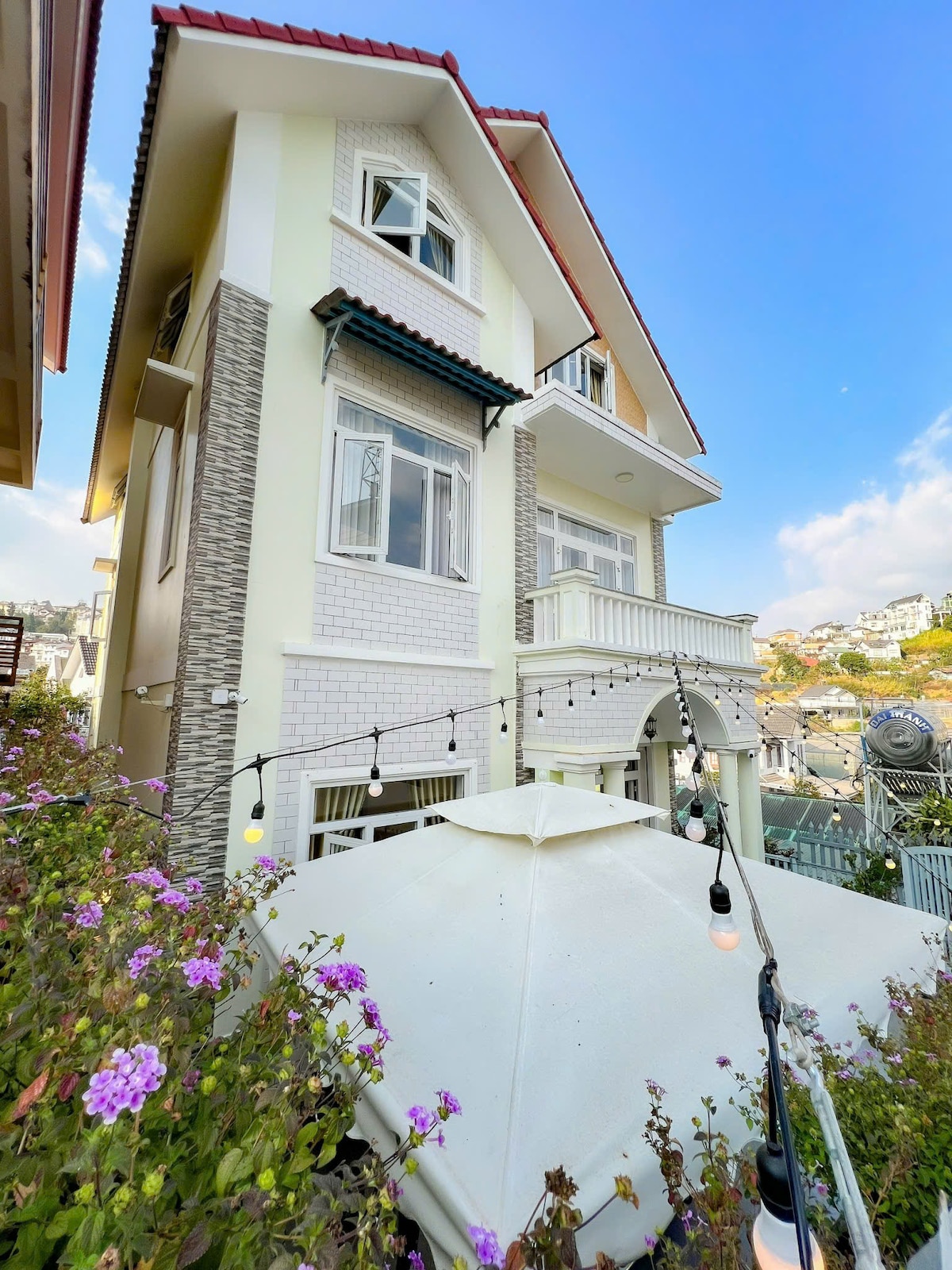 The exterior of the villa is showcased with a three-story structure featuring a combination of white and stone accents. A balcony with decorative railings is visible, surrounded by blooming flowers. Outdoor string lights add a welcoming touch to the inviting garden space below.