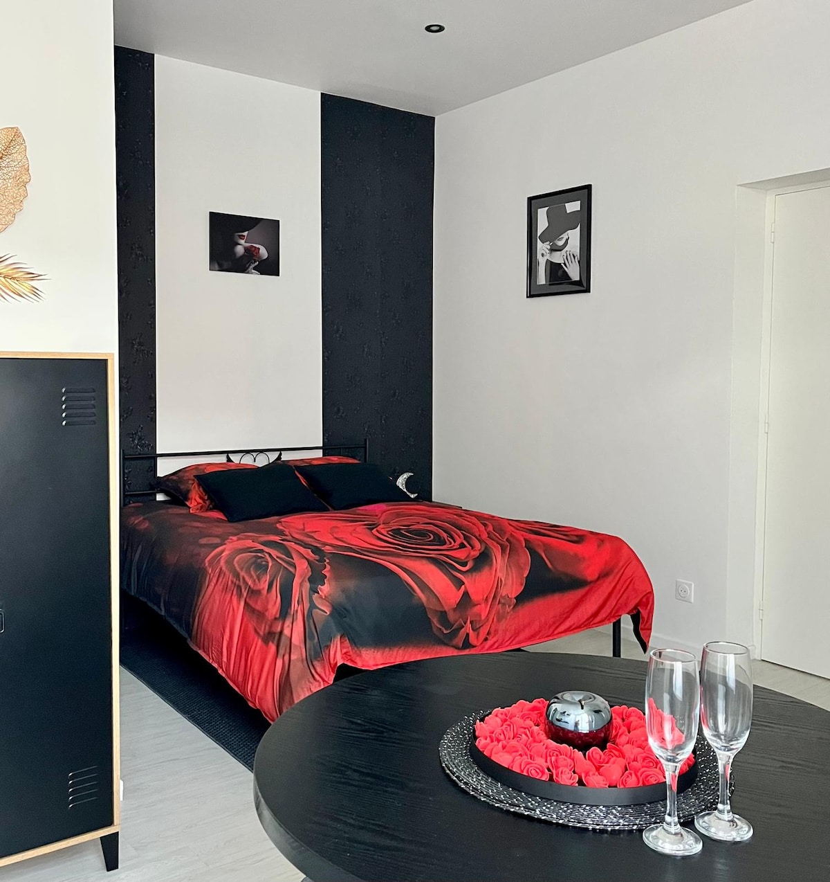 A cozy sleeping area features a bed adorned with red rose-patterned bedding. The walls are accented with black and white art prints. A small table with decorative roses and two champagne flutes is visible, contributing to the studio's romantic ambiance.