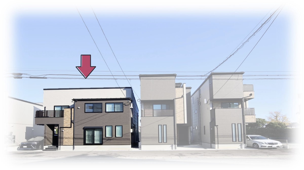 The two-story standalone house is situated between two neighboring structures. The exterior features a modern design with a combination of dark and light materials. Windows are visible, providing natural light. The entryway is marked by a balcony, while a clear blue sky serves as the backdrop.