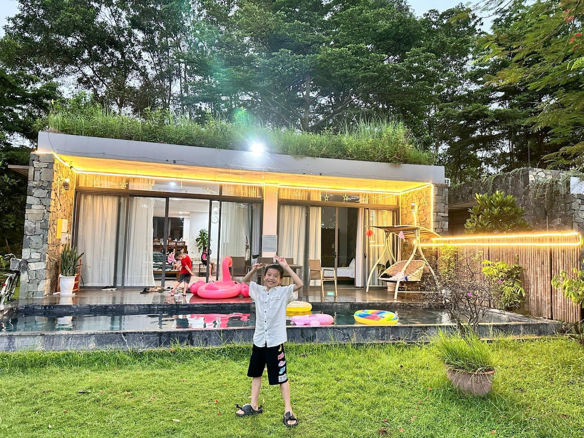 A modern villa is surrounded by lush greenery, featuring large glass doors that open to a private pool area. Brightly colored inflatable floaties are seen in the water. A child stands playfully in the foreground, capturing the lively atmosphere of the outdoor space.