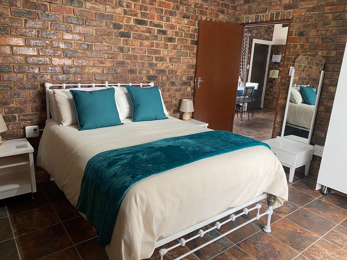 A well-appointed bedroom features a white bed with a soft cream coverlet and a teal throw. Two turquoise pillows sit at the head of the bed. Exposed brick walls complement the rustic ambiance, while a full-length mirror and bedside lamps are included in the inviting space.