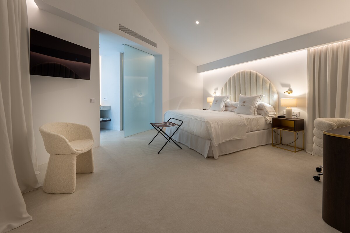 A modern bedroom features a spacious bed with a soft headboard and elegant bedding. A small folding table is positioned nearby, and accent lighting enhances the room's ambiance. A TV is mounted on the wall, with a translucent door leading to an adjoining private bathroom.