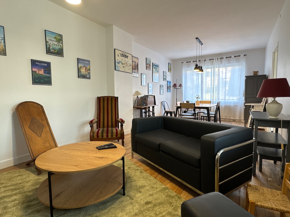 The living area features a modern black sofa facing a round wooden coffee table, set on a soft green rug. A striped armchair adds color, while the walls display various artworks. Natural light fills the room through large windows, with a dining area visible in the background.