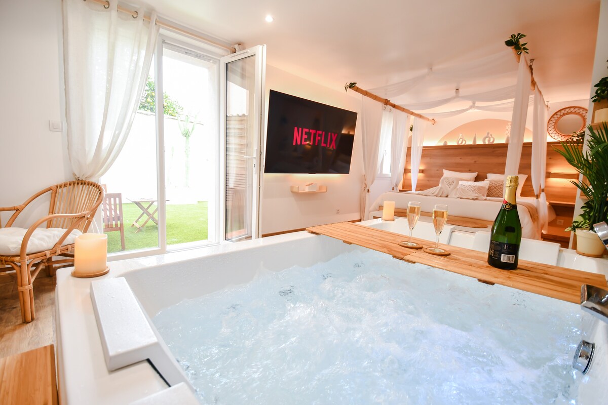 A spacious loft features a luxurious jacuzzi filled with bubbling water, complemented by a wooden surface that holds champagne flutes and a bottle of champagne. A large TV screen displays a streaming platform. Floor-to-ceiling windows provide access to an intimate garden.