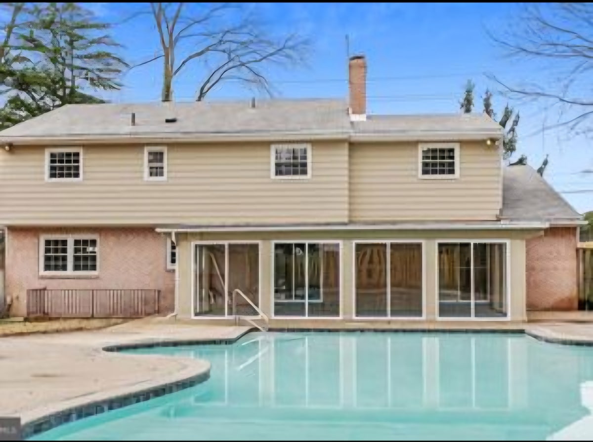 The back of the house features a spacious patio area surrounding an inviting pool. Large glass sliding doors provide an unobstructed view of the pool, and multiple windows enhance natural light. A lush green landscape is visible in the background, alongside a wooden fence for privacy.