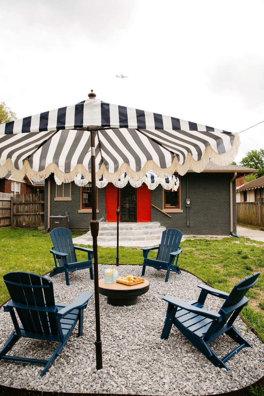 A cozy outdoor space features four blue Adirondack chairs arranged around a gravel area, shaded by a striped umbrella. A small table holds snacks and drinks, creating an inviting setting for relaxation. The backdrop includes a well-maintained fence and a view of the house.