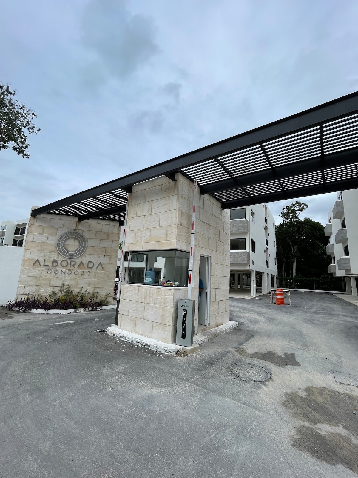 An entrance gate stands at the front of the condominium, featuring a modern design with a tiled facade. A security booth is visible, complemented by a roof structure. Surrounding greenery enhances the setting, with nearby buildings partially visible in the background.