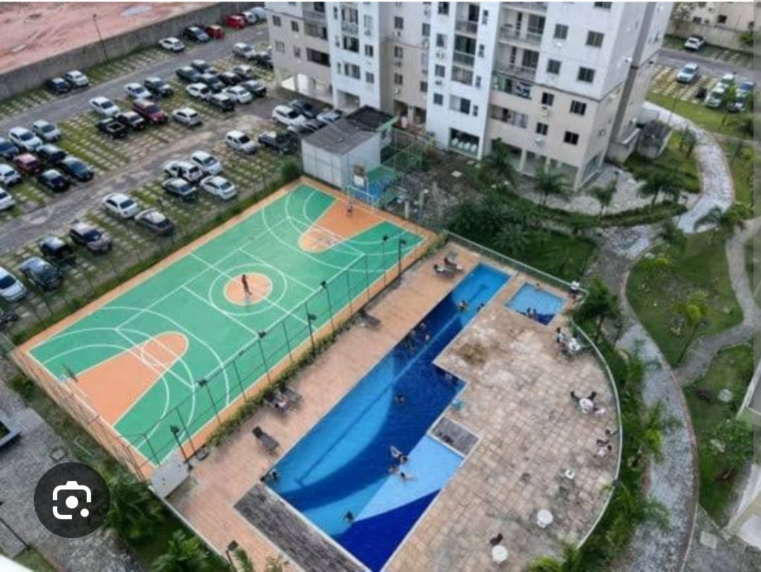 An aerial view captures a sports court and a swimming pool surrounded by greenery. The vibrant green and orange basketball and tennis courts are in use, while the pool area features lounge chairs for relaxation. Nearby vehicles are parked in a spacious lot.