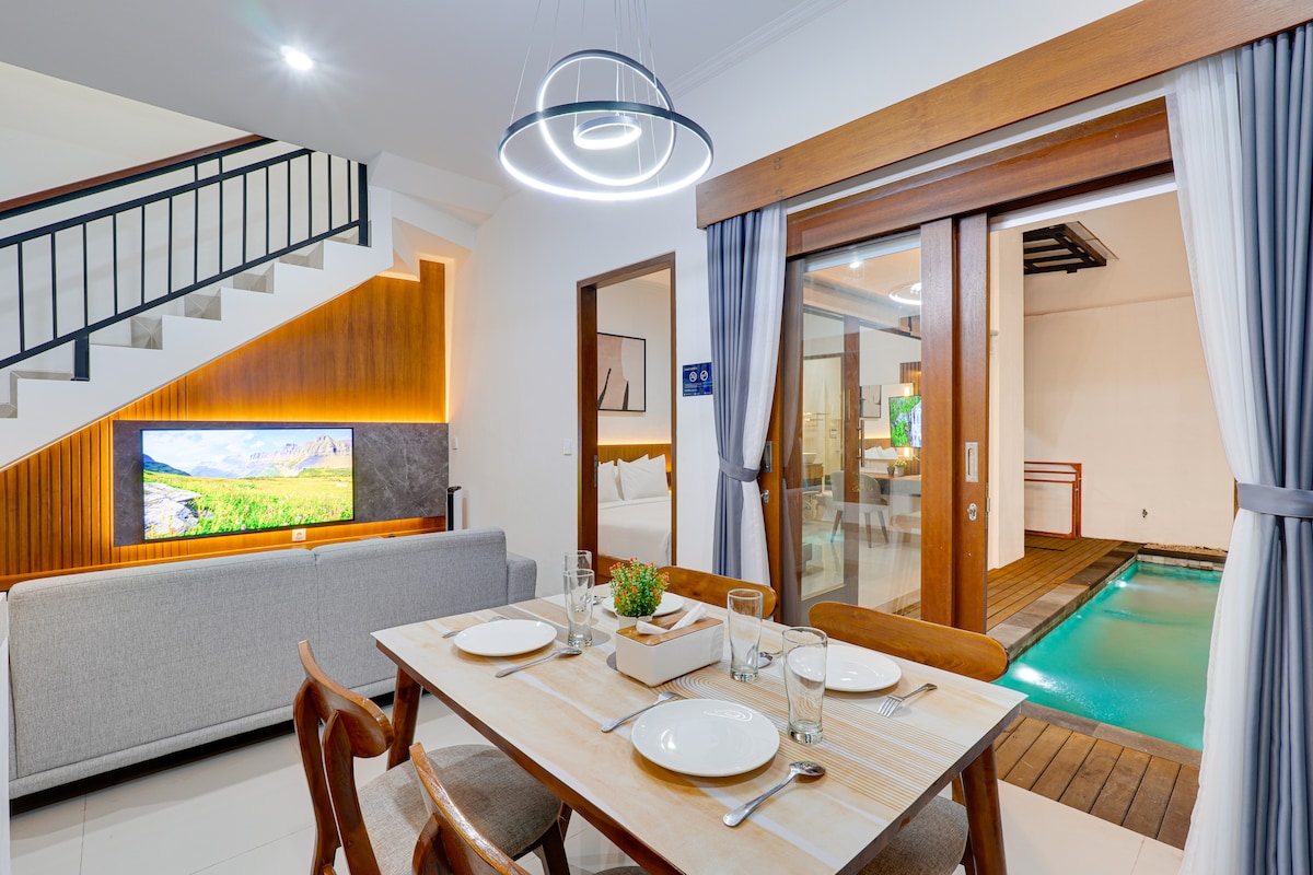 An inviting living and dining area features a wooden dining table set for four, with glasses and plates arranged neatly. A large flat-screen TV is mounted on the wall, while glass doors provide a view of the private pool area. Natural light fills the space.