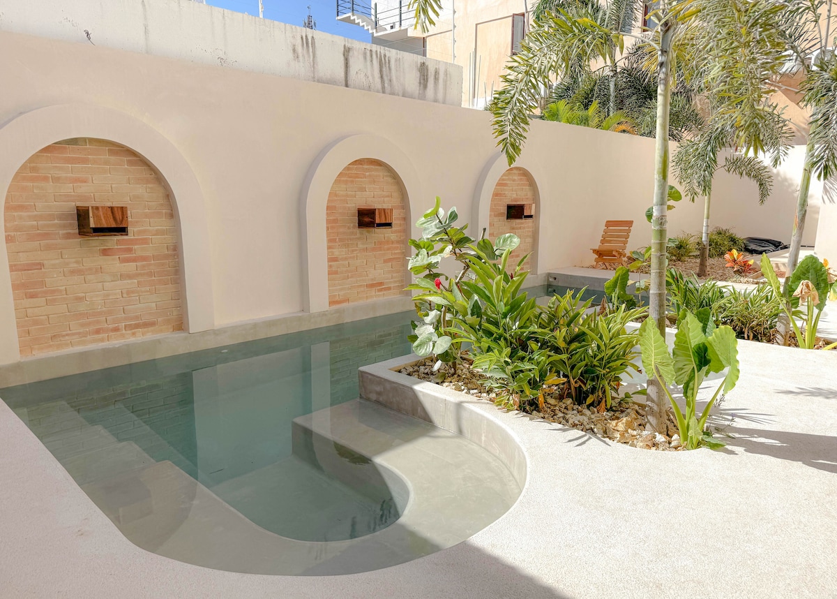 A serene pool area is surrounded by lush greenery and tropical plants. Arched niches with wooden accents provide a decorative touch against a smooth white wall. A comfortable wooden lounge chair is positioned nearby, inviting relaxation in a peaceful environment.