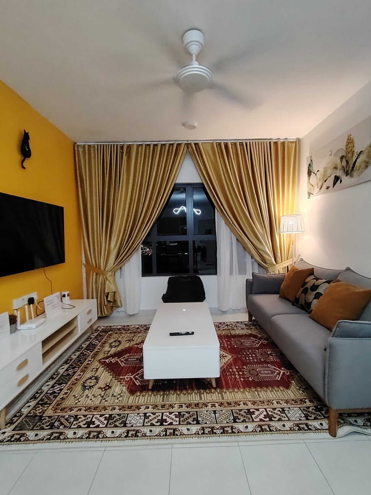 A living area features a grey sofa adorned with orange and black decorative pillows. A white coffee table is centered on an ornate area rug. Yellow curtains frame a large window, complemented by wall art and a television on a minimalist cabinet.