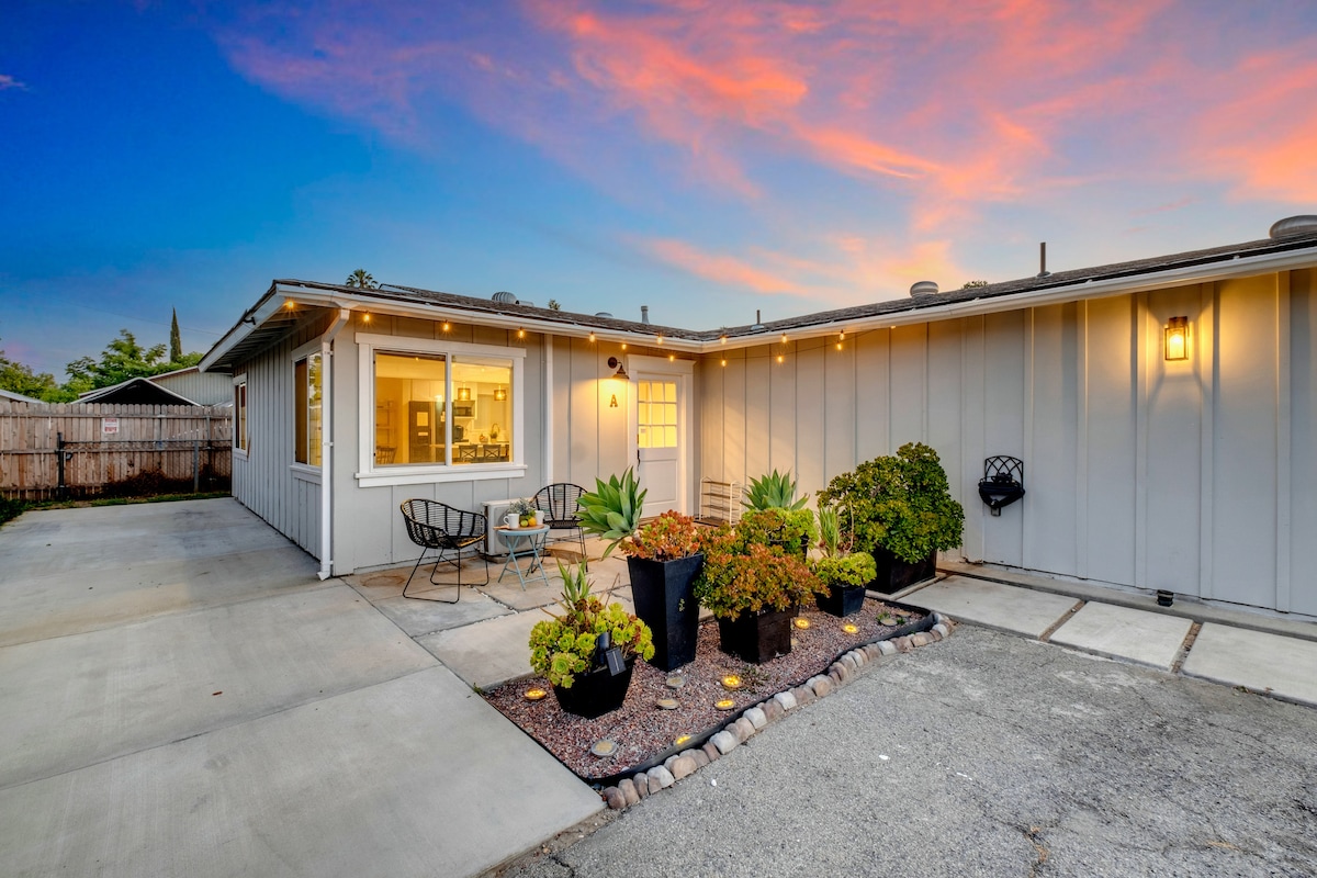 An inviting entrance showcases a landscaped area with vibrant plants and flowers, surrounded by decorative stones. A driveway leads to the house, where a large window offers a view into the bright interior. String lights enhance the outdoor ambiance against a colorful sunset sky.