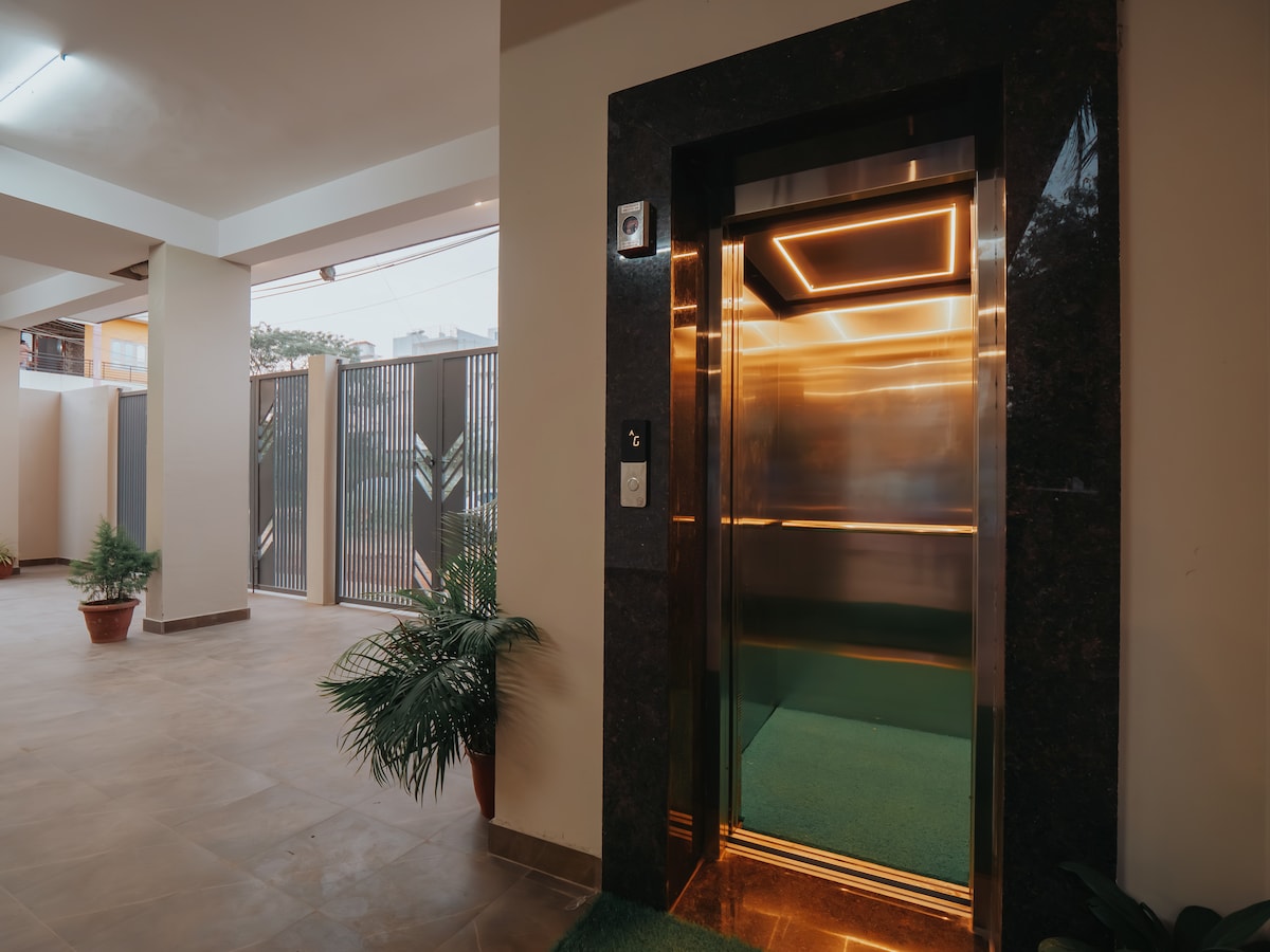 An elevator entry is framed by sleek marble accents, positioned in a well-lit corridor. The surrounding area features potted plants, while a gated entrance is visible, enhancing accessibility for guests. The clean lines and modern design create a welcoming environment.