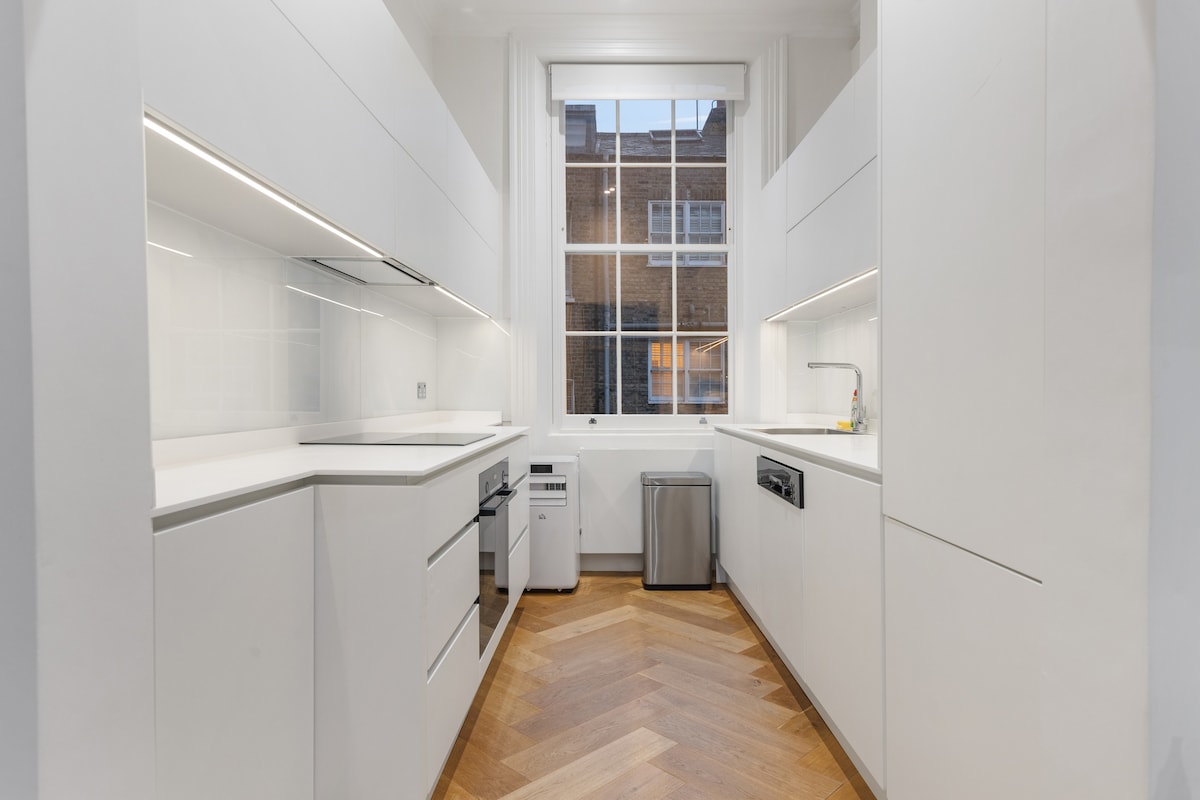 A modern kitchen features sleek white cabinetry and clean lines. High-quality Siemens appliances are present, alongside ample counter space for meal preparation. A large window allows natural light to illuminate the area, enhancing the open and airy atmosphere.