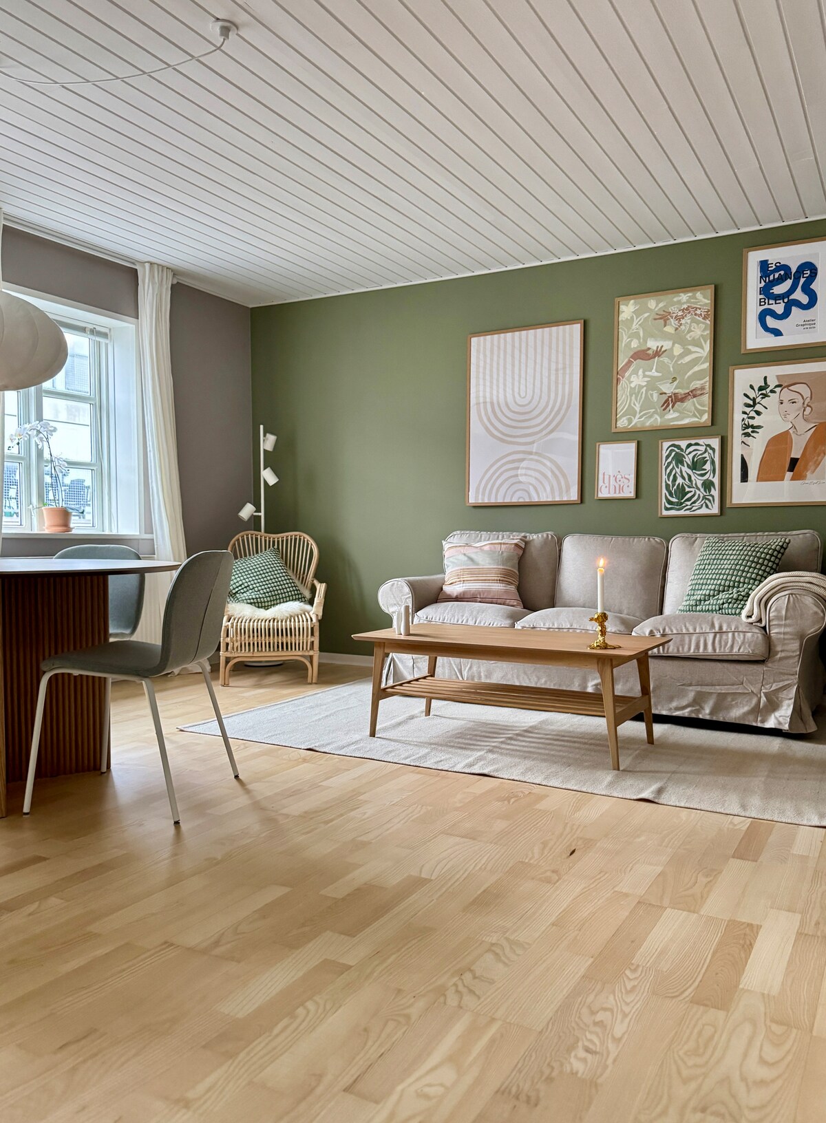 A cozy living area features a light-colored sofa adorned with soft cushions and a decorative throw. A wooden coffee table rests on a neutral rug, complemented by a dining table and modern chairs nearby. Artwork is displayed on the green wall, creating a harmonious ambiance.