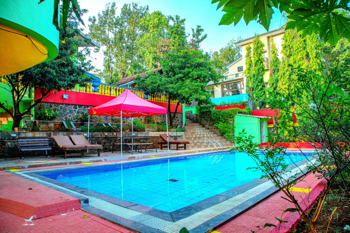 A swimming pool is surrounded by vibrant greenery and colorful structures. Sun loungers are positioned nearby, offering a space for relaxation. A red canopy provides shade over part of the pool area, with steps leading up to a landscaped elevation in the background.