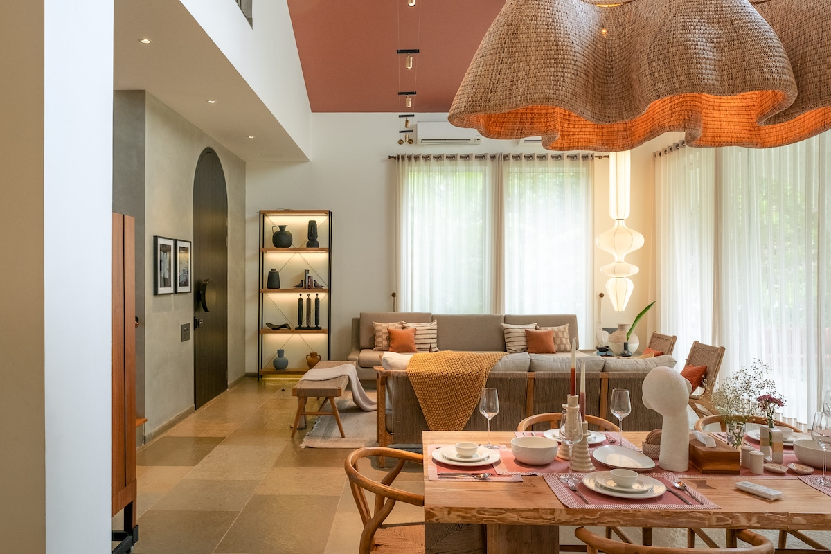 An open living area features a large sectional sofa adorned with pillows, complemented by a wooden dining table set for a meal. Natural light pours in through floor-to-ceiling windows, while softly textured lighting fixtures hang above, enhancing the space's warm and inviting feel.