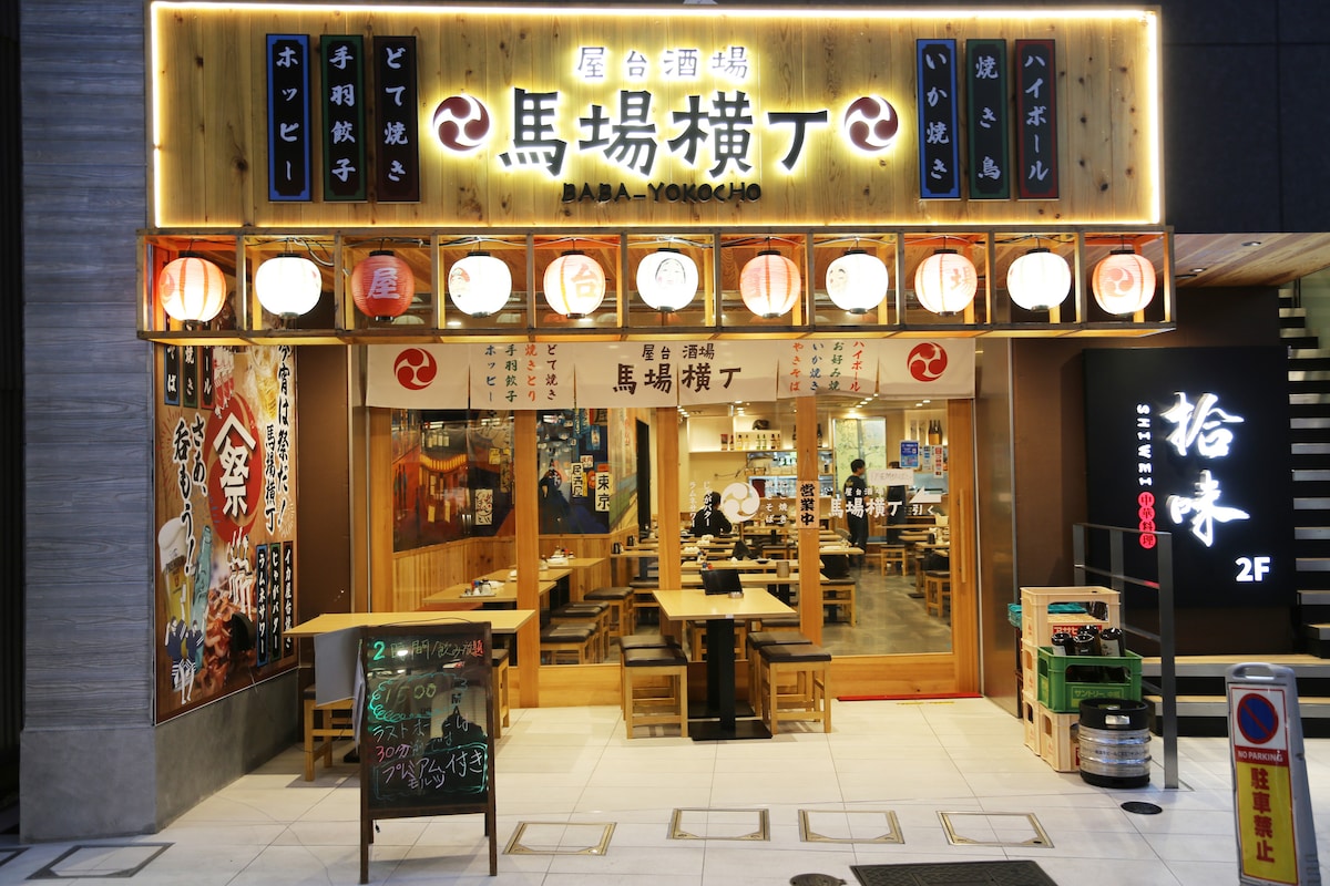A vibrant izakaya features wooden elements and hanging lanterns, creating a lively atmosphere. Outdoor seating is available under a decorative awning, while a chalkboard displays menu offerings. The open interior reveals a spacious dining area, encouraging a social dining experience.