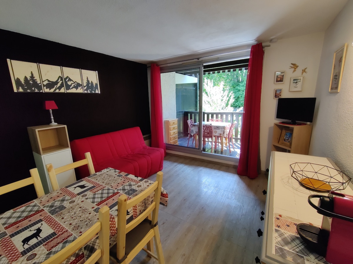 The living area features a red sofa bed, a dining table with wooden chairs, and a TV unit. Large glass doors open to a balcony, allowing natural light and views of the outdoors. Bright red curtains frame the windows, contrasting with the dark accent wall.