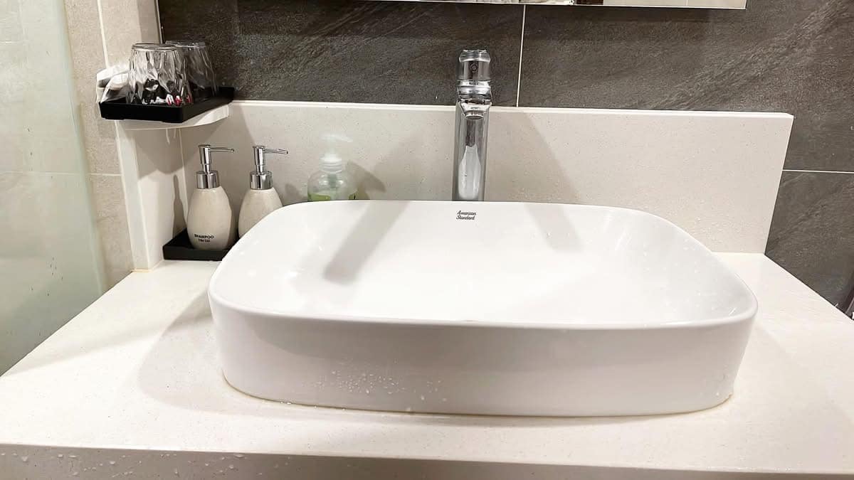 A modern bathroom sink is presented on a smooth countertop. The square basin is white, accompanied by a sleek faucet positioned above. Adjacent shelves hold drinking glasses and toiletries, with two bottles visible. The overall design emphasizes cleanliness and functionality.