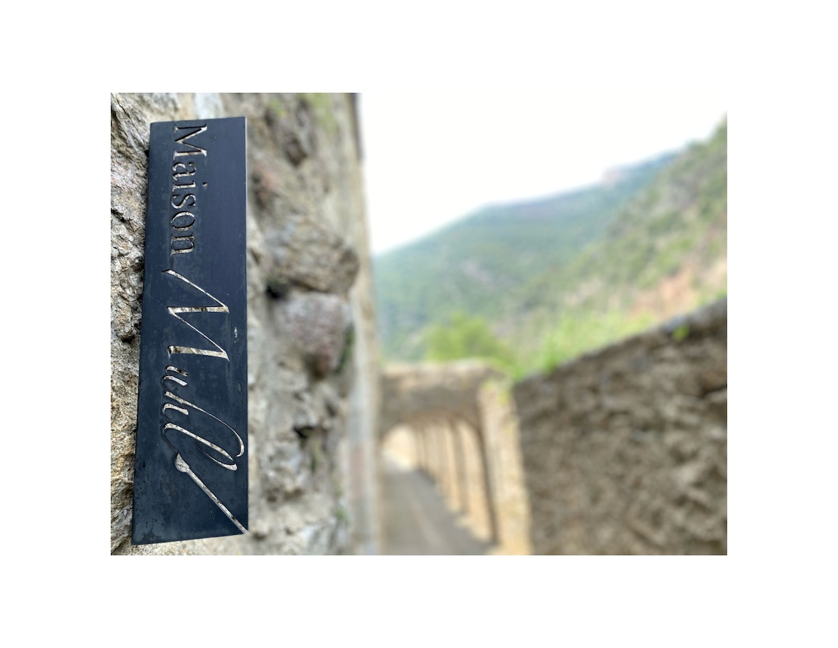 A metal sign displaying 'Maison Mulh' is affixed to a stone wall, surrounded by a historic village atmosphere. The background features a soft-focus mountain landscape, further enhancing the visual appeal of the ancient architecture along the pathway.