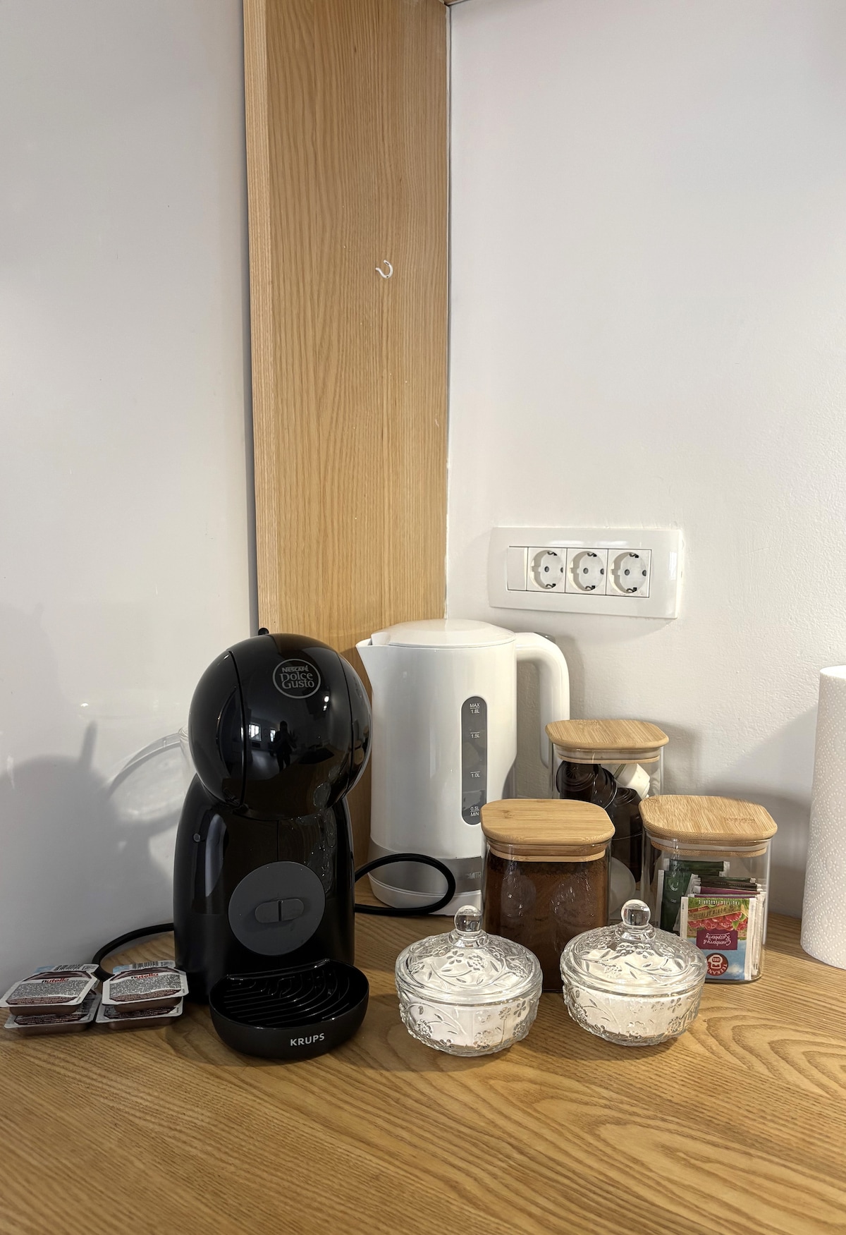 A well-equipped kitchen corner is presented with a sleek black coffee machine, a kettle, and assorted canisters. Transparent containers hold coffee and tea supplies, while decorative jars provide a touch of elegance. A wooden countertop complements the appliances, creating a functional and organized space.