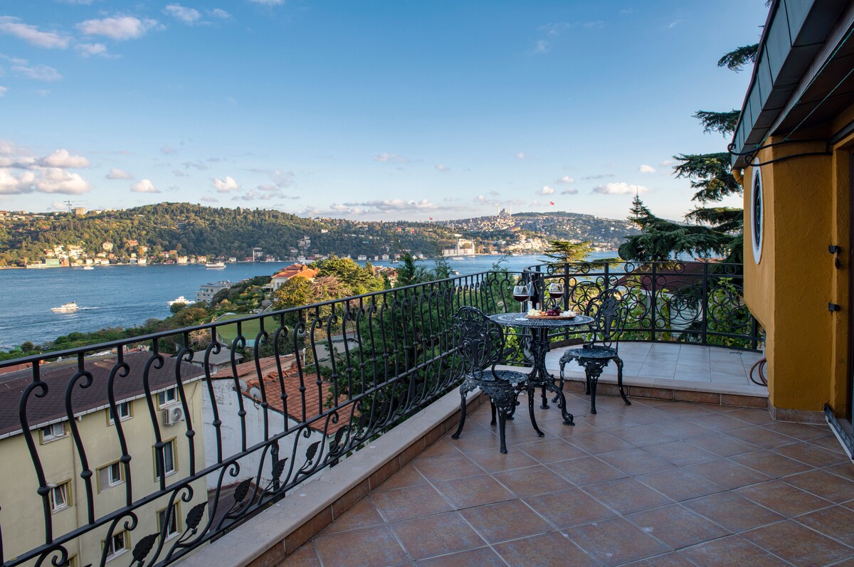A balcony with ornate metal furniture offers a stunning view of the Bosphorus and surrounding landscape. Sunny skies and occasional clouds add to the scenic backdrop. The terrace floor is tiled, and the railing features intricate designs, enhancing the open space's visual appeal.