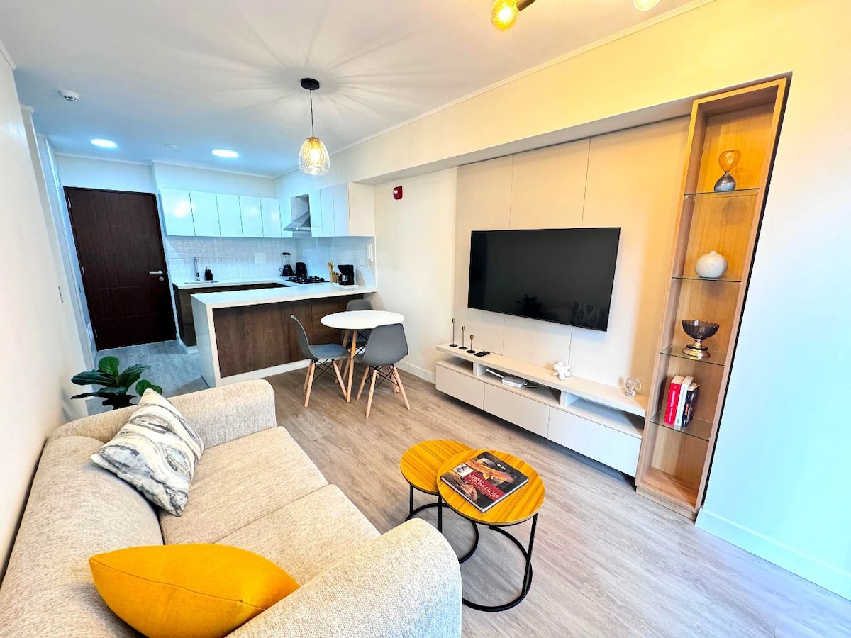 A modern living area is visible, featuring a light gray sofa with a decorative yellow pillow. A flat-screen television is mounted on the wall opposite a compact entertainment unit. A dining table for four is positioned beside a sleek kitchen with white cabinetry.