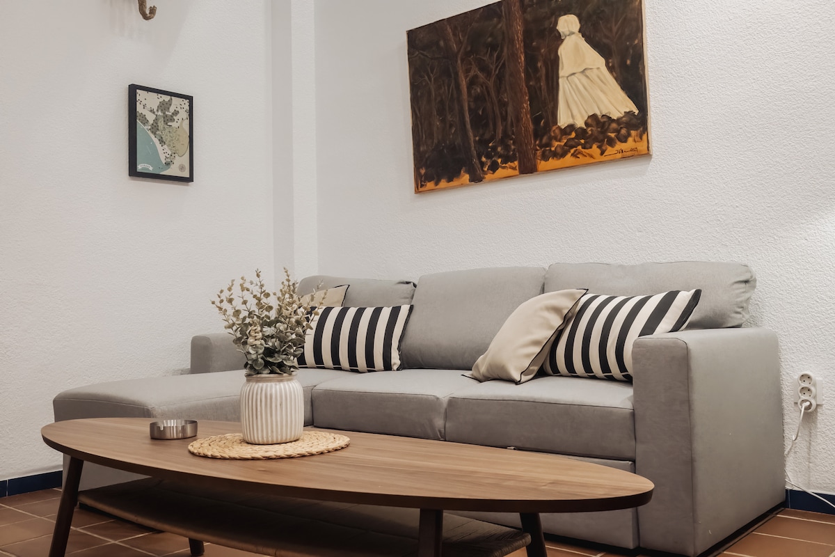 A comfortable gray sectional sofa is positioned against a white wall, featuring decorative cushions in black and white stripes. A wooden coffee table with a round woven placemat sits in front, accompanied by a small vase holding dried flowers and a candle.