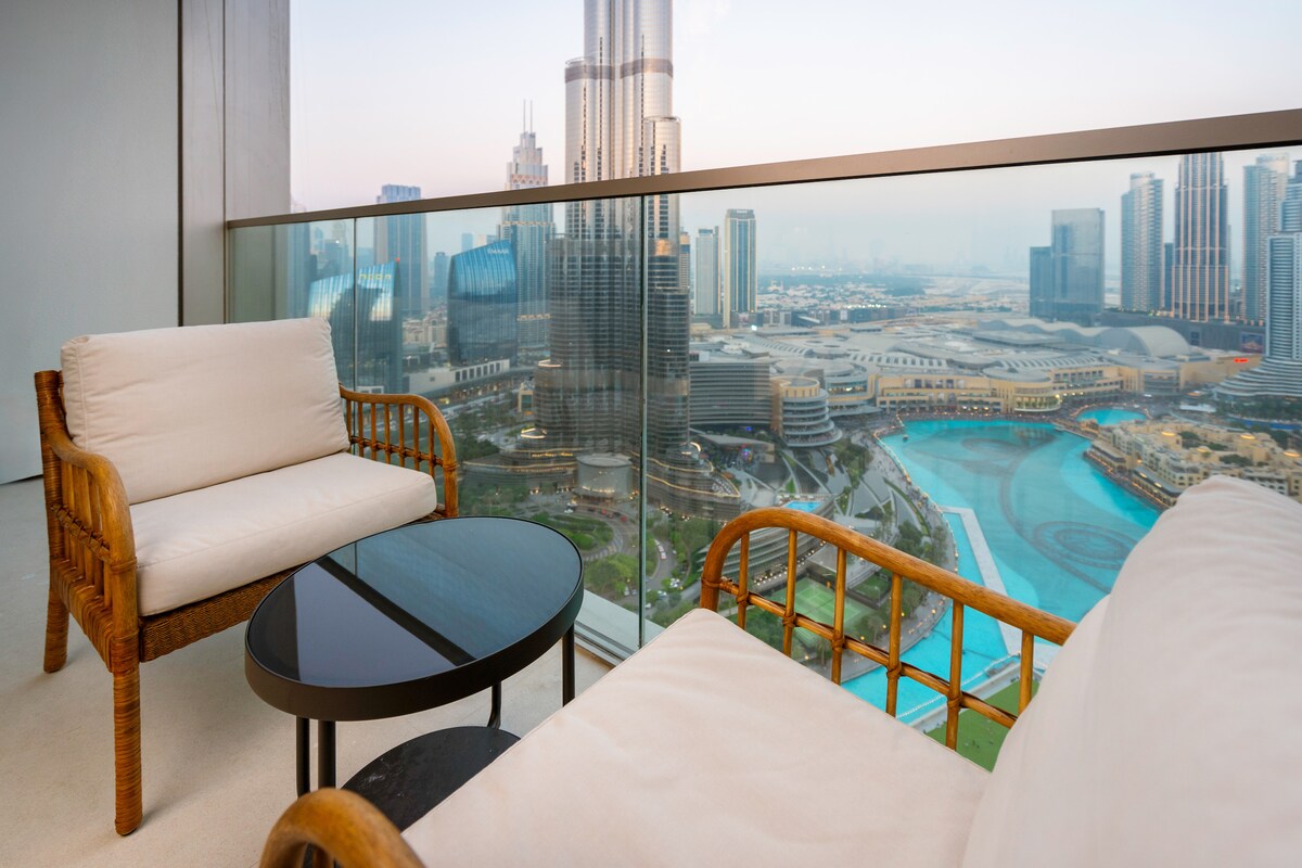 A spacious balcony features cozy seating arrangements with two armchairs and a round table. The view overlooks the iconic Burj Khalifa and the Dubai Fountain, with the vibrant cityscape providing a stunning backdrop during sunset.