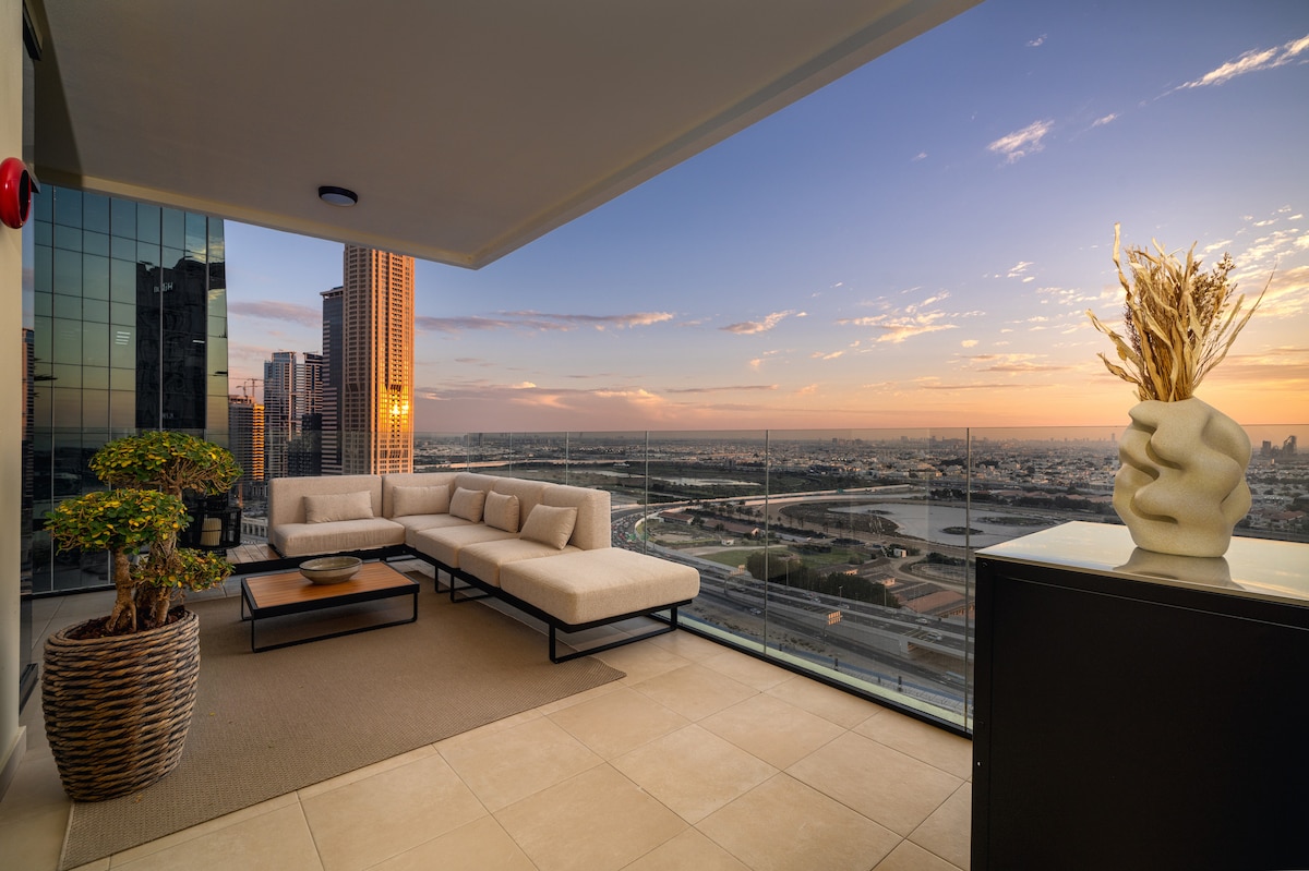 A spacious terrace features modern lounge seating with low-profile cushions positioned for optimal sunset views. The glass railing provides an unobstructed panorama of the city skyline and canal, while a decorative plant adds a touch of greenery to the setting.
