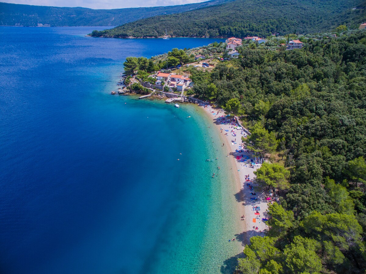 Aerial view of a coastal area featuring a sandy beach lined with umbrellas and beachgoers. Clear blue waters blend into deeper shades, surrounded by lush greenery and rocky outcrops. The charming village is visible in the background, nestled among the trees along the shoreline.