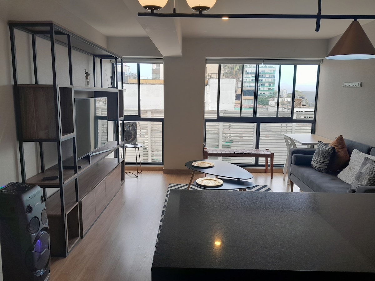 A modern living area is presented with large windows providing natural light and offering views of the surrounding city. A sleek entertainment unit features a flat-screen TV, complemented by a comfortable sofa and two minimalist coffee tables. A dining area is visible in the background.