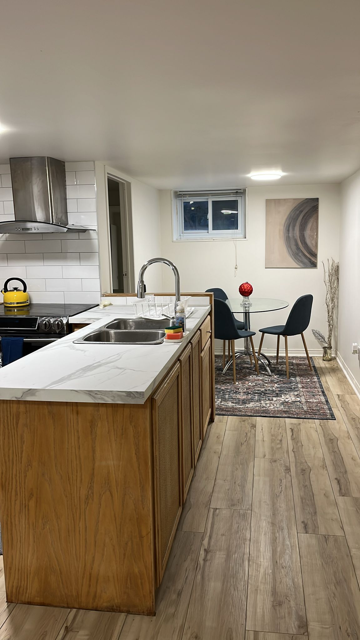 An open-concept kitchen and dining area features a marble-topped island and a stainless steel sink. A round dining table with four chairs sits nearby, accompanied by a decorative area rug. Natural light enters through a small window, enhancing the bright and modern space.