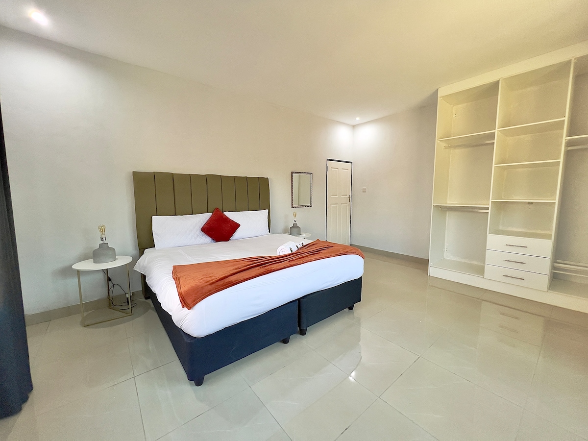 A spacious bedroom is highlighted by a comfortable bed with crisp white linens and an accent pillow. A decorative orange throw adds a touch of color. Nearby, a nightstand with a lamp complements the minimalist style of the room, featuring light-colored walls and tile flooring.