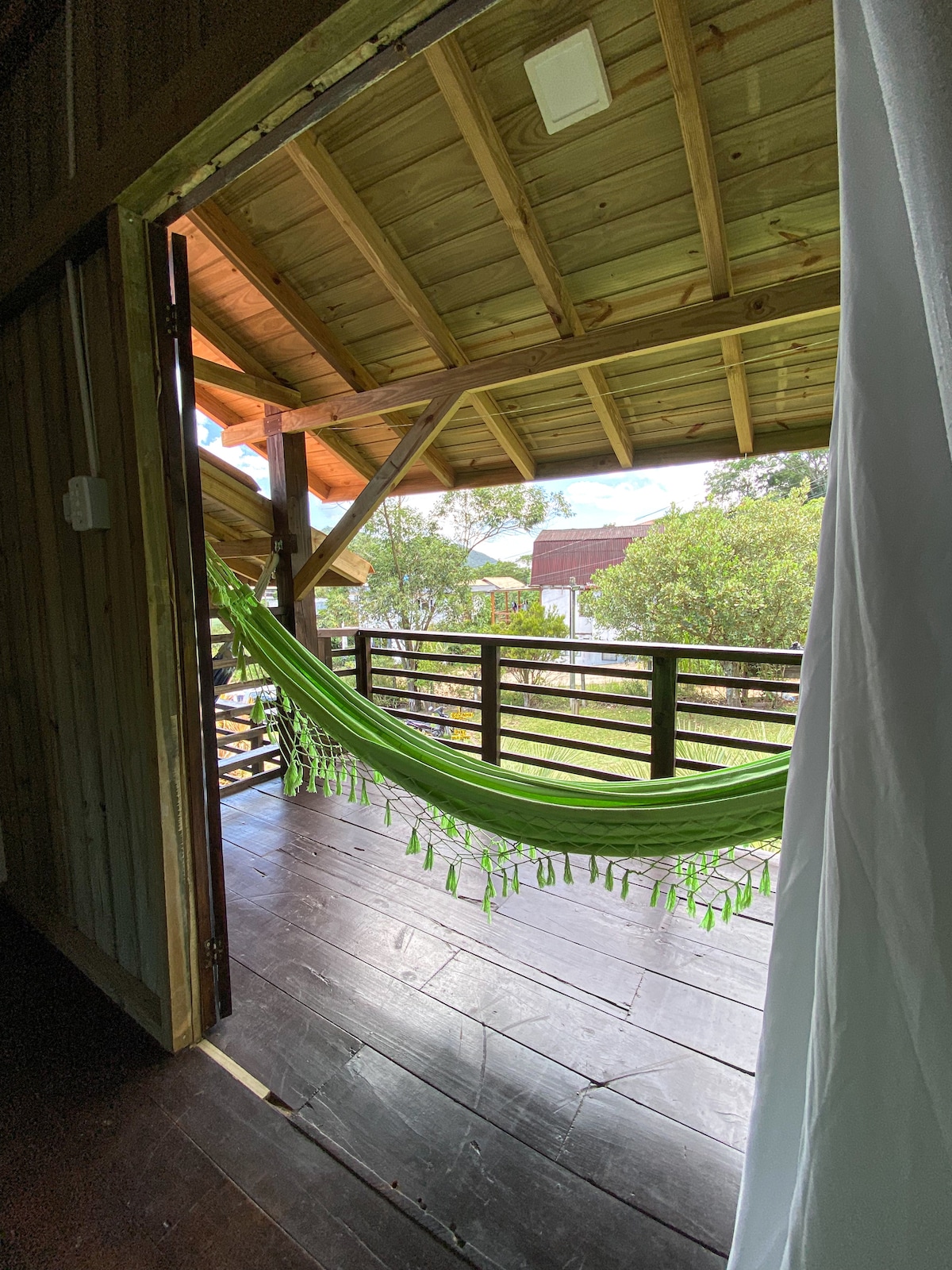 A bright and airy balcony is visible, featuring a green hammock suspended between wooden posts. The open space reveals a view of the surrounding nature, with ample light filtering through the structure's wooden beams.