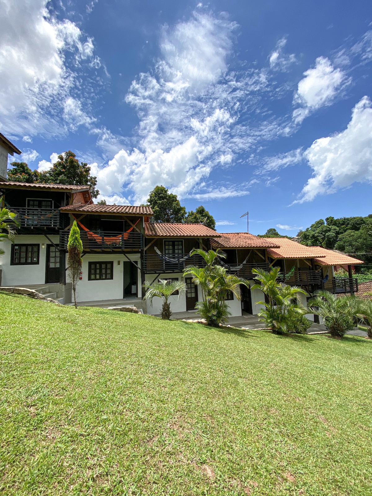 A two-story building is depicted, featuring multiple bungalows with charming architecture and terracotta roofs. The grassy area in front is landscaped with palm trees, and a backdrop of lush greenery is visible under a bright blue sky with scattered clouds.
