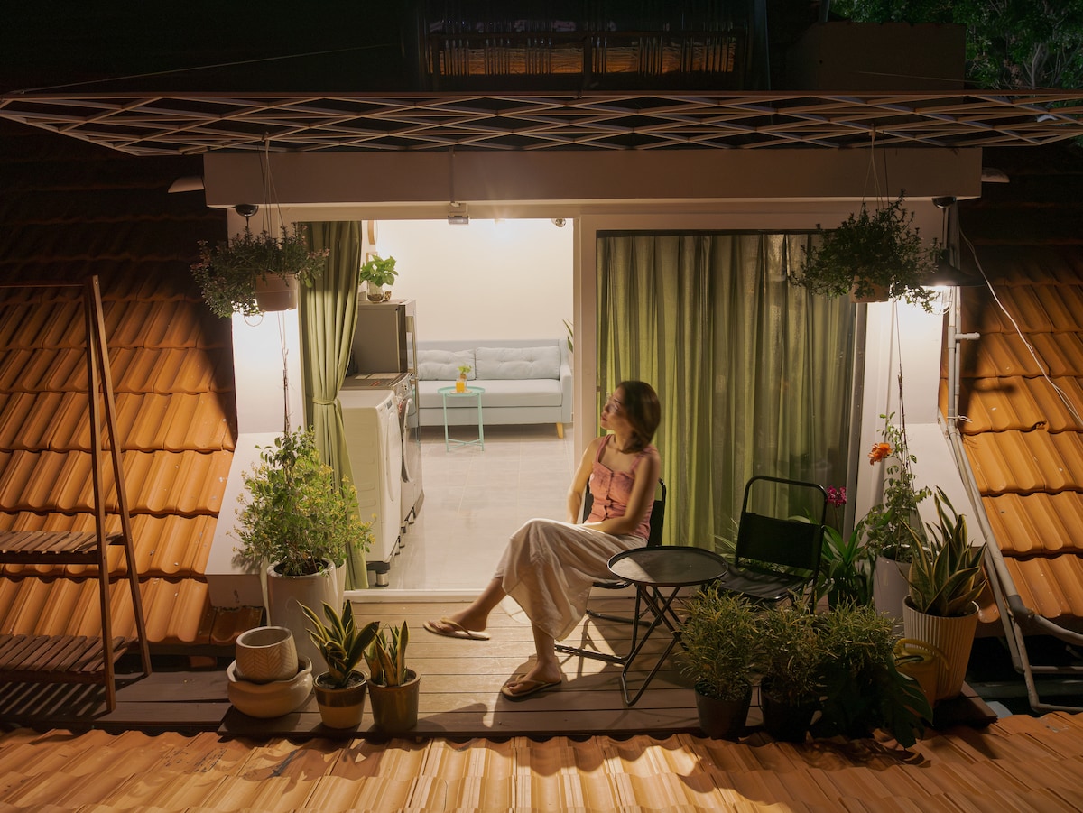 A serene outdoor balcony is observed, featuring potted plants and a small table with chairs. Soft lighting illuminates the area, while lush green curtains drape the windows behind, contrasting with the warm terracotta roof tiles. A figure relaxes peacefully, enjoying the ambiance.
