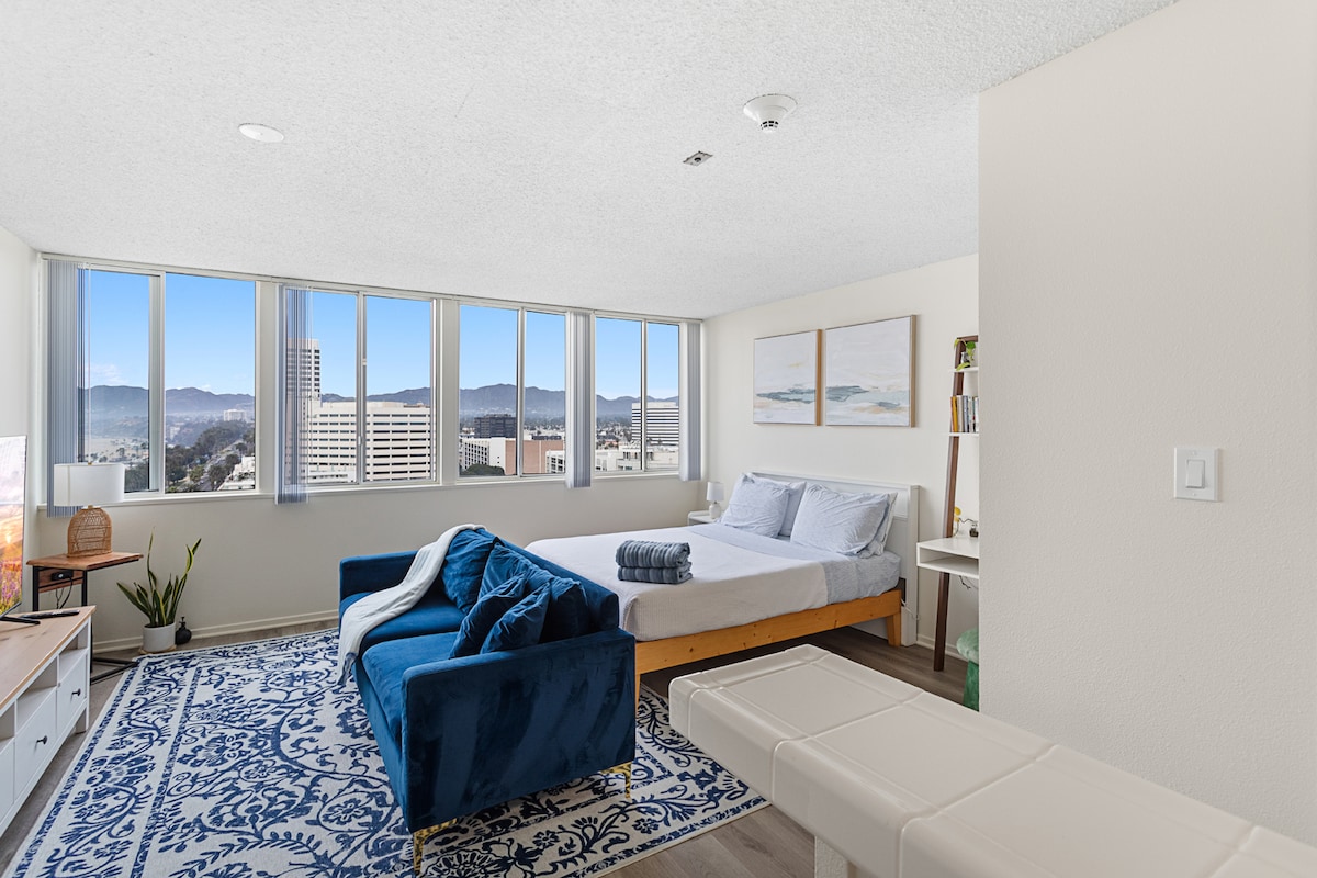 A bright studio space features large windows offering city views. The room contains a spacious bed with light bedding and a cozy blue sofa. Elegant artwork is displayed on the walls above the bed, and a small table is positioned nearby, complemented by a decorative plant.