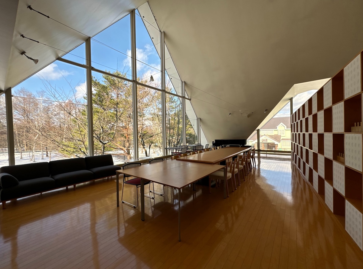 A spacious common area features a large wooden table surrounded by dining chairs, complemented by a comfortable dark sofa. Floor-to-ceiling windows allow natural light to fill the space, offering views of the surrounding trees and landscape.