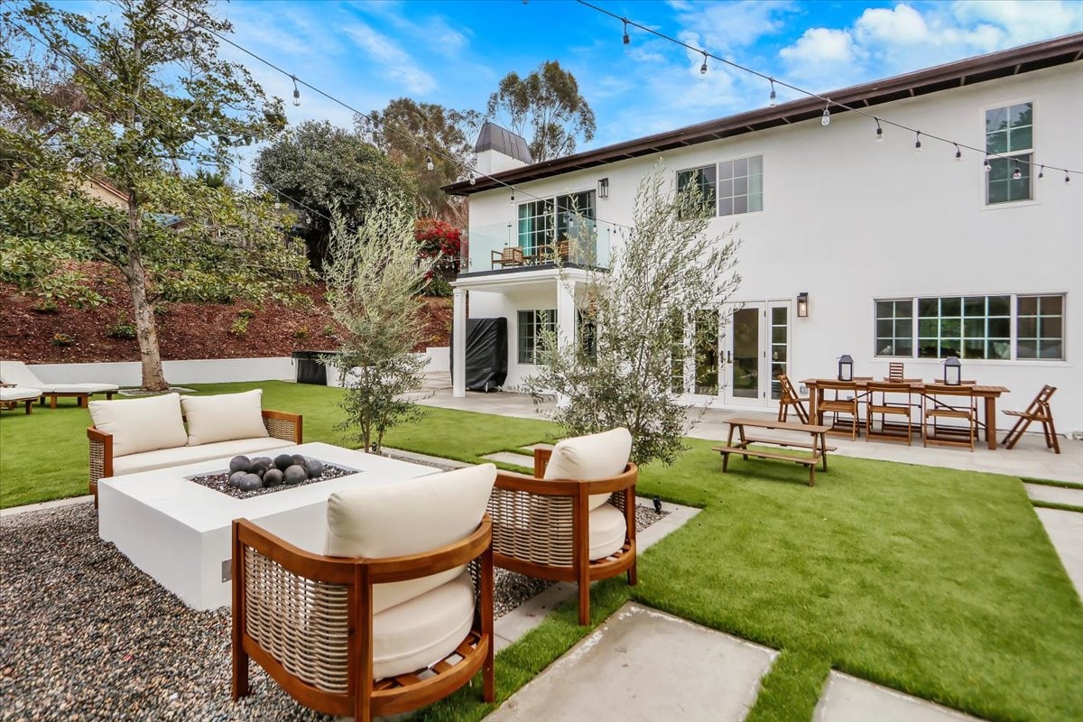 An outdoor space features a fire pit surrounded by four comfortable chairs. Nearby, a long dining table accommodates several guests. Lush green grass and decorative gravel enhance the patio, while string lights are seen overhead, adding a soft glow to the serene environment.
