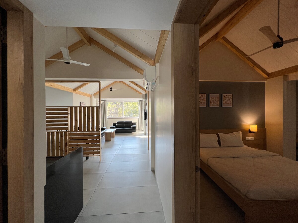 An interior view showcases a spacious room with high wooden ceilings and ample natural light. A cozy bed is positioned in one area, while a separate living space features a sofa and a large TV. Neutral colors create a serene atmosphere throughout.